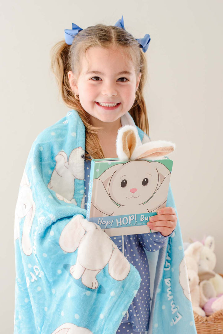 Little girl in pigtails smiling while holding the "Hop! Hop! Bunny!" board book, wrapped in a Caden Lane personalized blue bunny-print blanket with embroidered name visible.