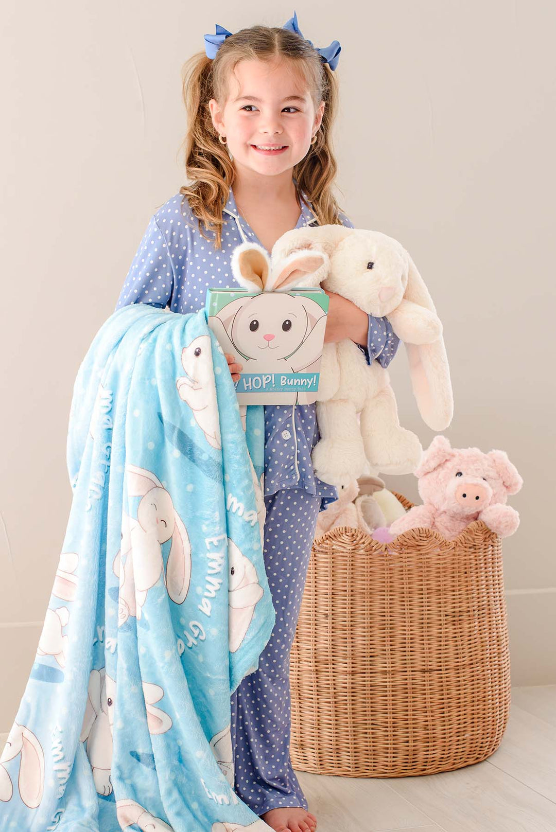 Smiling little girl in pigtails holding a plush bunny toy and the "Hop! Hop! Bunny!" board book, standing beside a draped Caden Lane personalized blue bunny-print blanket and a basket of stuffed animals.