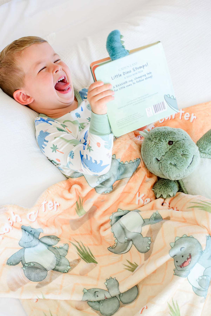 Toddler boy in Caden Lane dinosaur pajamas lying in bed laughing while holding the "Little Dino Stomps!" board book, wrapped in a Caden Lane personalized orange dinosaur-print blanket with a plush green dinosaur toy beside him.