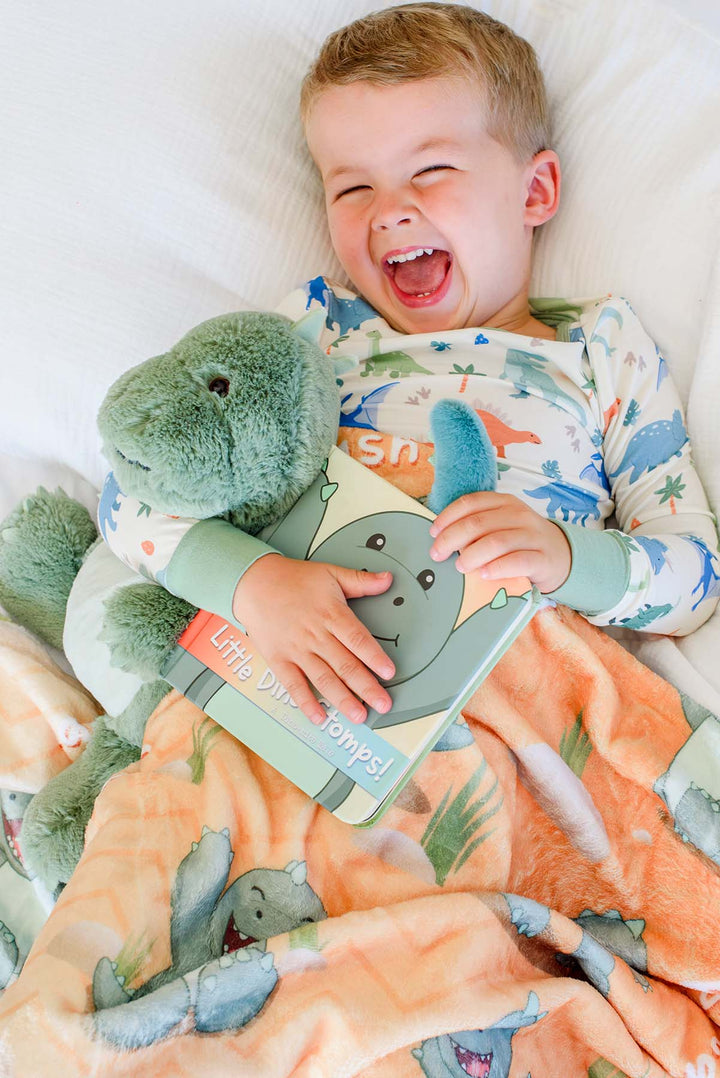 Close-up of a laughing toddler in Caden Lane dinosaur pajamas hugging a plush green dinosaur toy and the "Little Dino Stomps!" board book, lying on a Caden Lane personalized orange dinosaur-print blanket