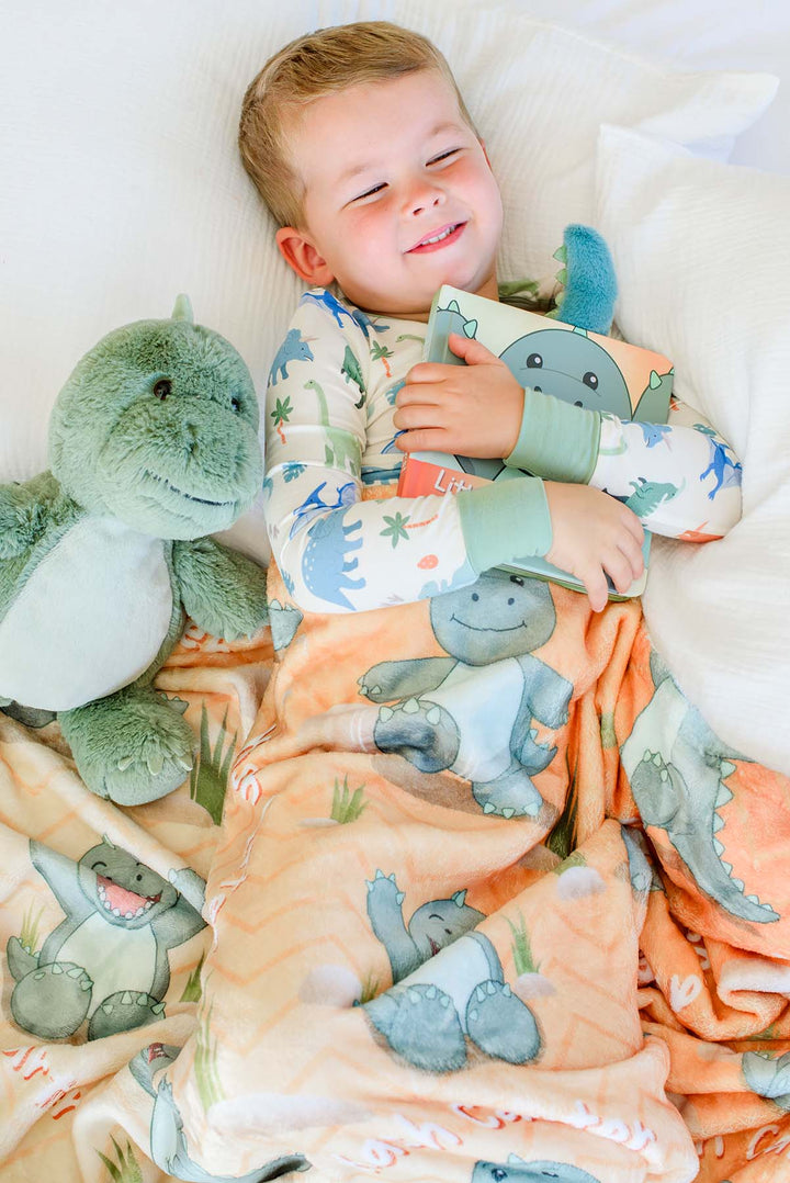 Smiling toddler lying in bed hugging a plush green dinosaur and the "Little Dino Stomps!" board book, tucked under a Caden Lane personalized orange dinosaur-print blanket with embroidered name visible