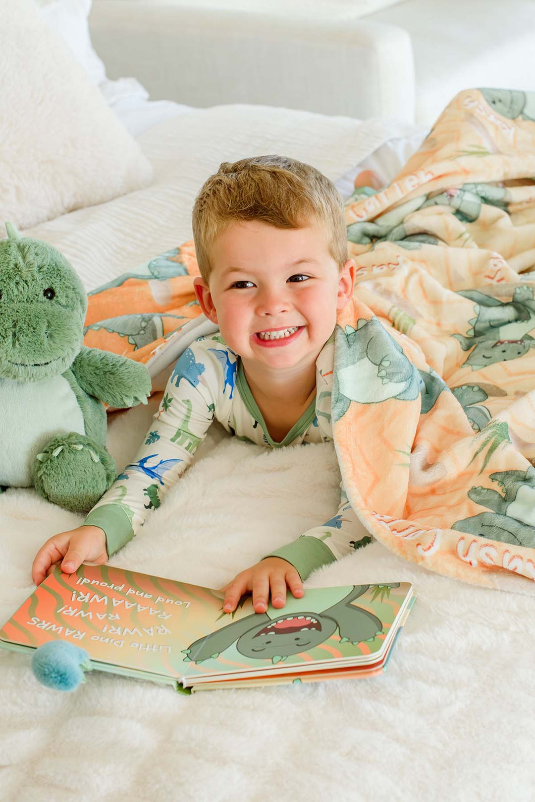 Toddler boy grinning while peeking out from under a Caden Lane personalized orange dinosaur-print blanket, with a plush green dinosaur beside him and an open "Little Dino Stomps!" board book in front of him