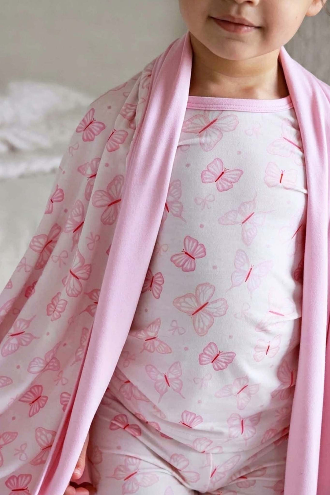 Two Piece Pajama Set in Blooming Butterflies print - child wearing the butterfly print pajama top draped with a pink bamboo blanket, showing the pajama fabric close-up