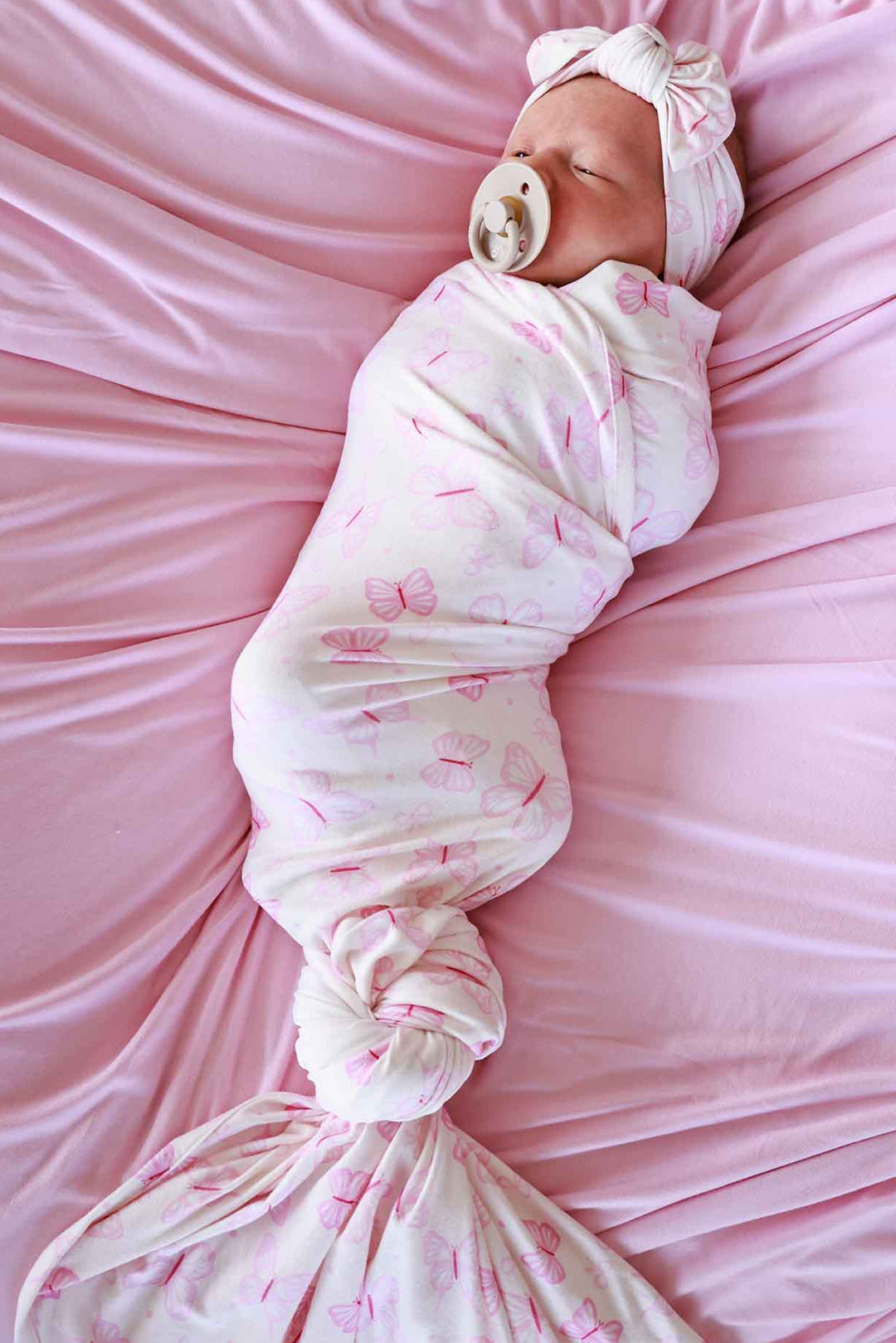 Oversized Swaddle Blanket in Blooming Butterflies print - newborn tightly swaddled in the pink butterfly-print blanket on pink satin, wearing a matching white bow headband