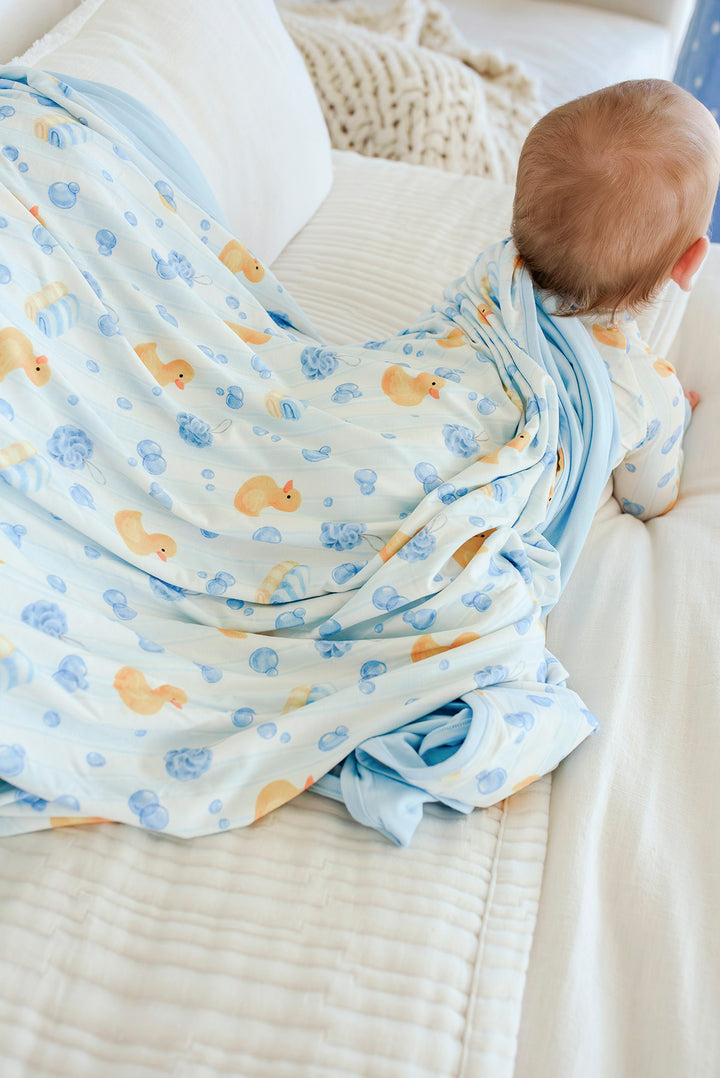 Baby lying on blue Bubble Duckies double-sided bamboo blanket showing printed duck side and solid blue reverse