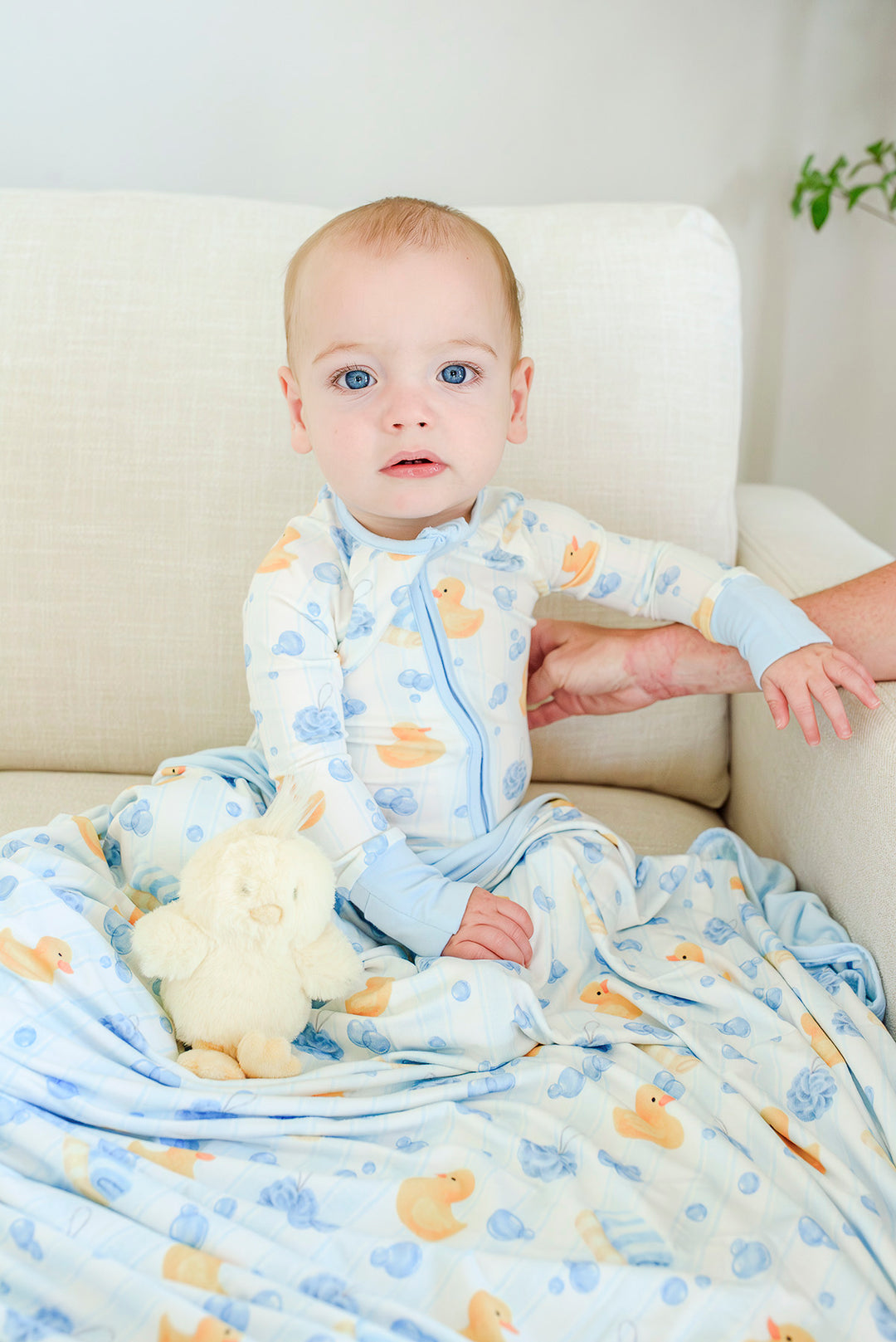 Baby sitting up in blue Bubble Duckies convertible zip romper with rubber duck stuffed toy and matching blanket