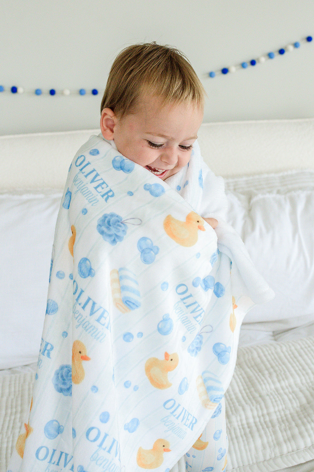 Young boy wrapped in blue Bubble Duckies personalized kids blanket with yellow rubber duck and blue bubble print featuring custom name