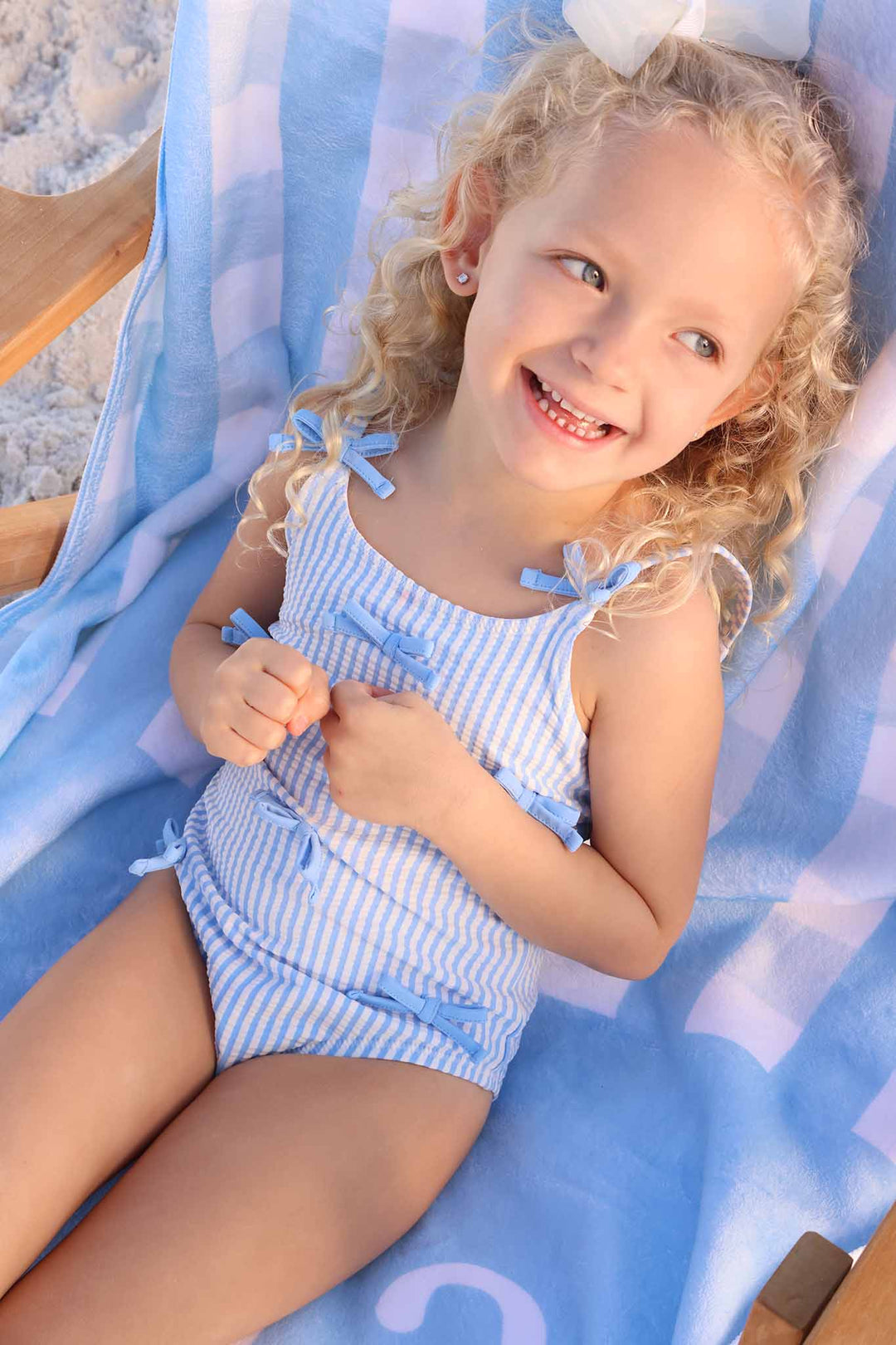 girl laying on lounge chair in blue seersucker one piece swimsuit with blue bows on the front 