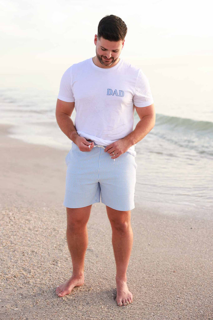 dad standing on the beach in blue seersucker men's swim trunks and dad tee