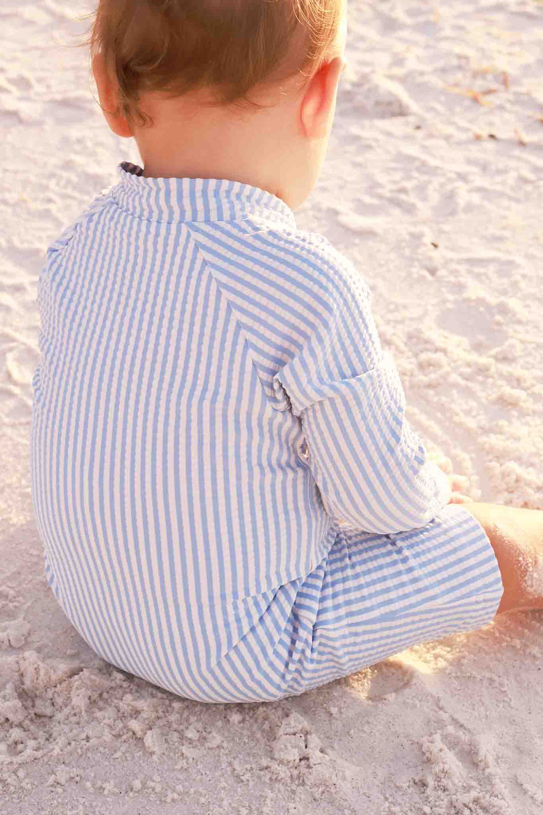 boy sitting on the beach in blue seersucker romper shortie