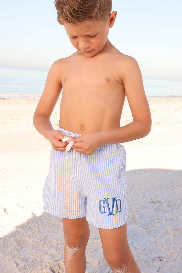 boy at the beach tying swim trunks in blue seersucker print with monogram embroidery 