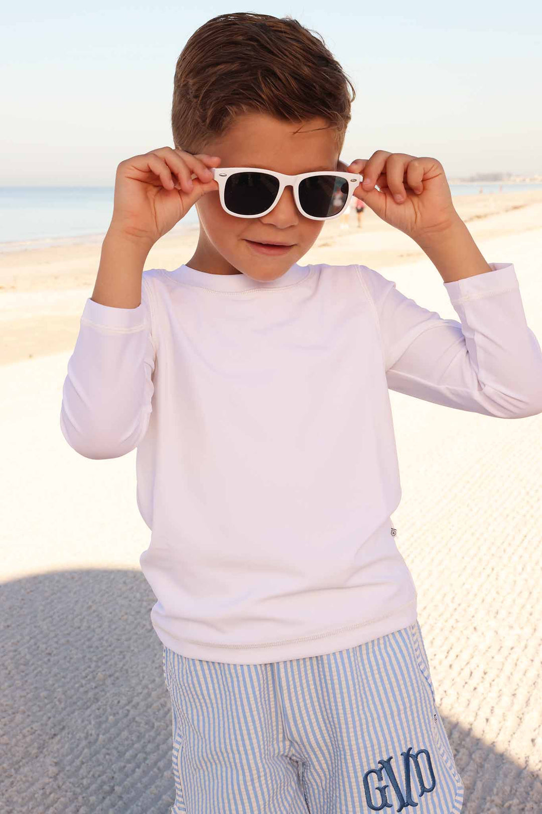 boy on beach wearing long sleeve rash guard shirt in white with seersucker shorts with blue monogram