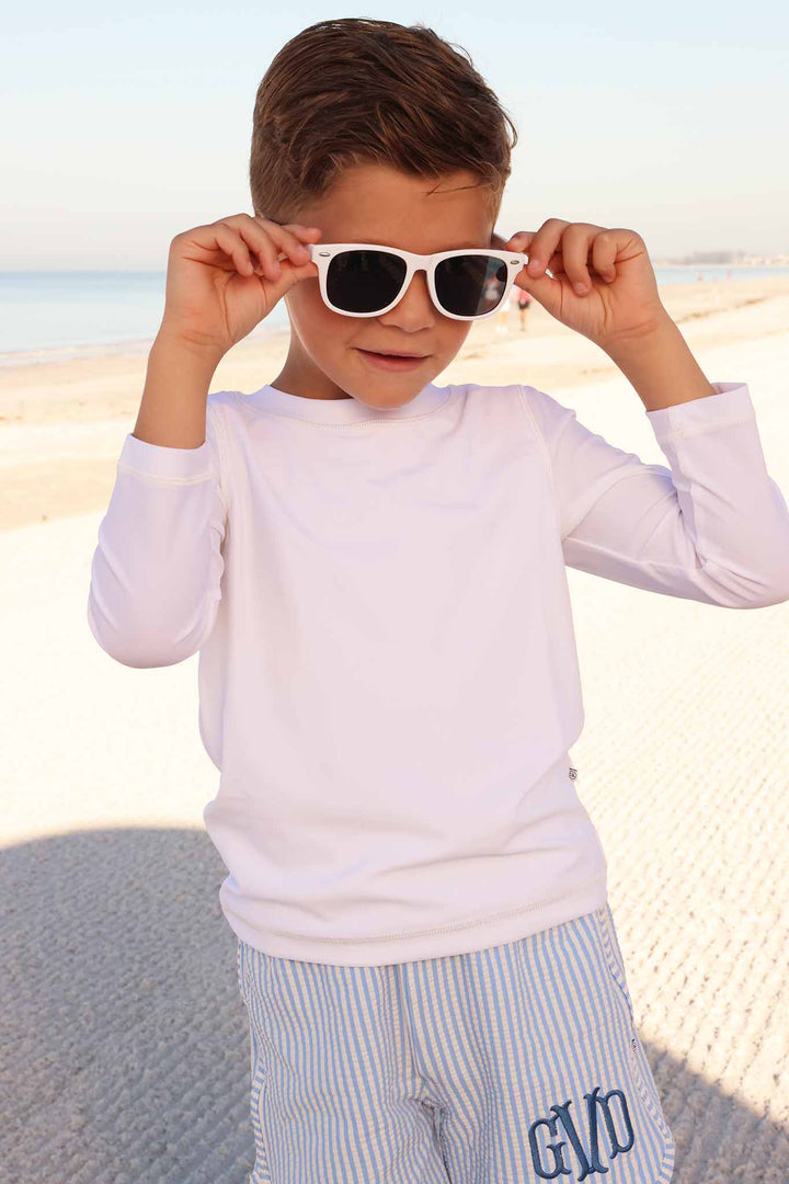 boy on beach wearing long sleeve rash guard shirt in white with seersucker shorts with blue monogram