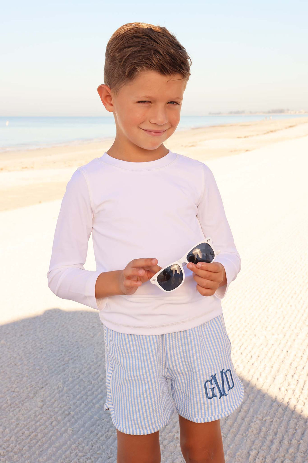 boy standing on beach in long sleeve rash guard shirt in white with blue seersucker swim trunks holding white sunglasses