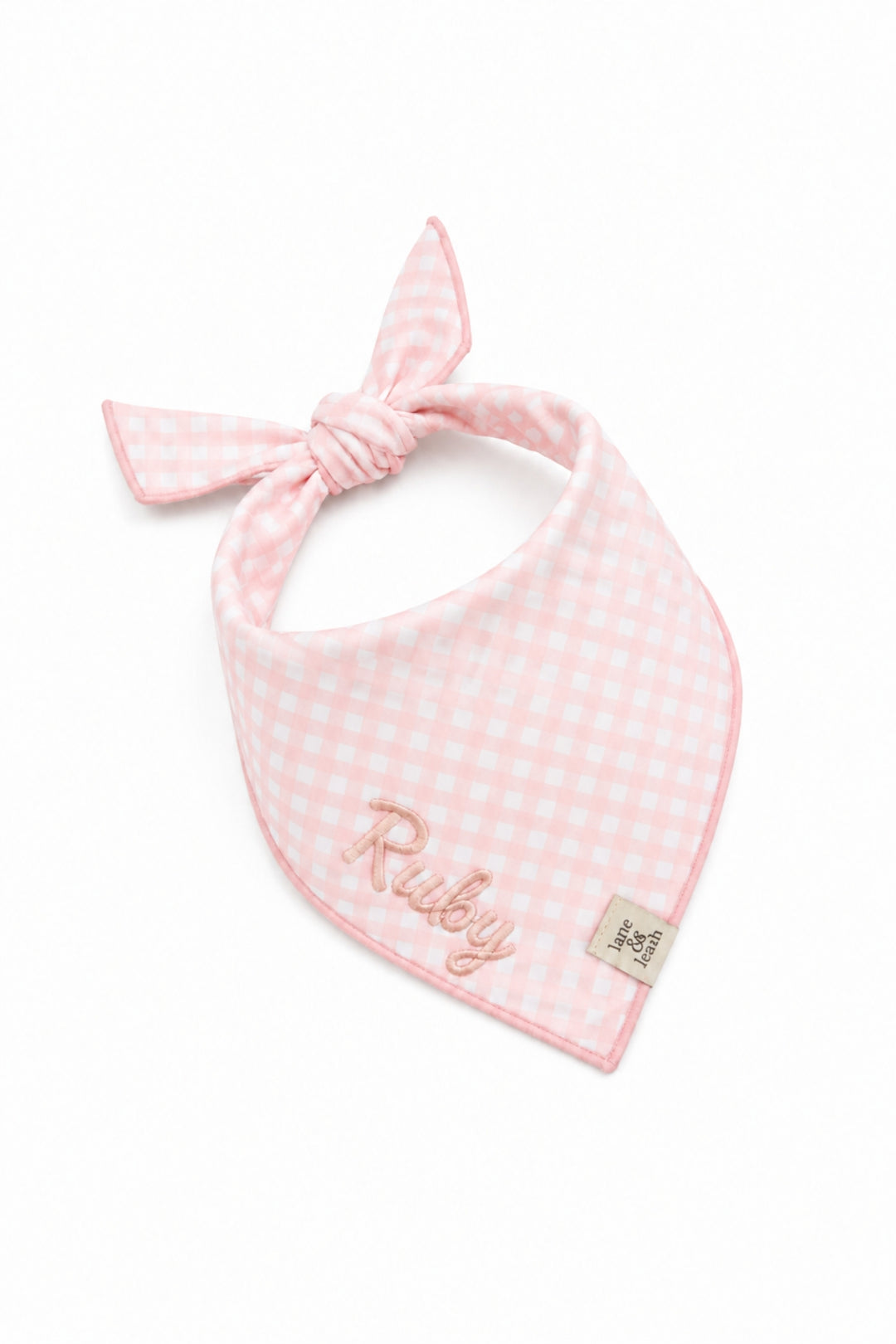 A pink and white gingham Lane & Leash dog bandana bib with a tie-neck design, featuring the name 'Ruby' embroidered in pink script and a small brand tag on the bottom right corner, set against a plain white background.