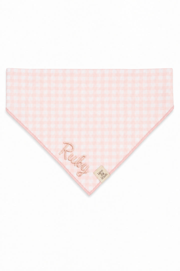 A Lane & Leash light pink and white gingham check pet bandana with 'Ruby' embroidered in rose gold script and a small beige brand tag, shown on a plain white background.