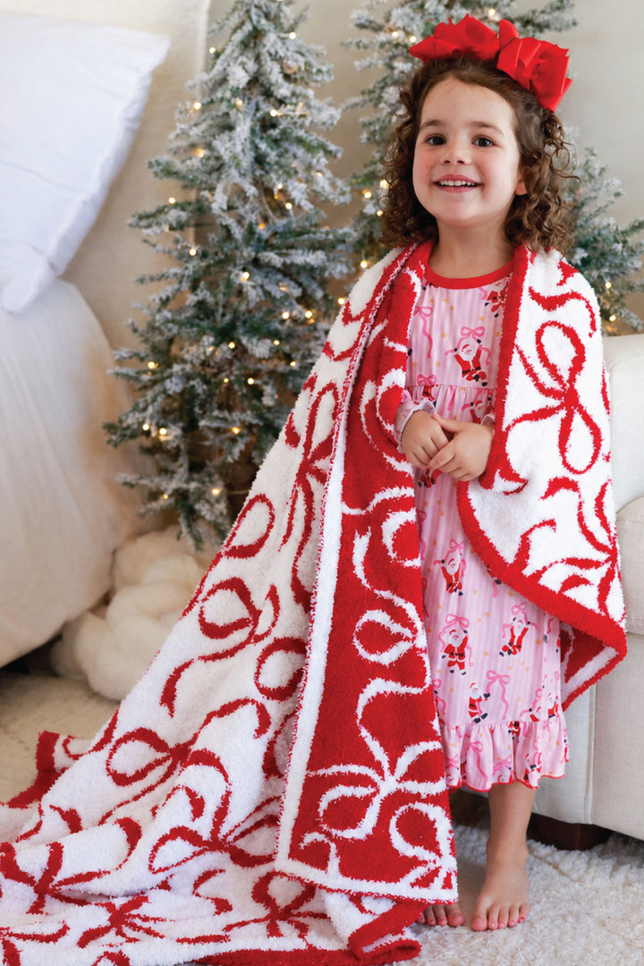 Caden Lane red and white bow print sherpa blanket draped over a smiling toddler girl in a Santa-print nightgown, standing in front of a decorated Christmas tree