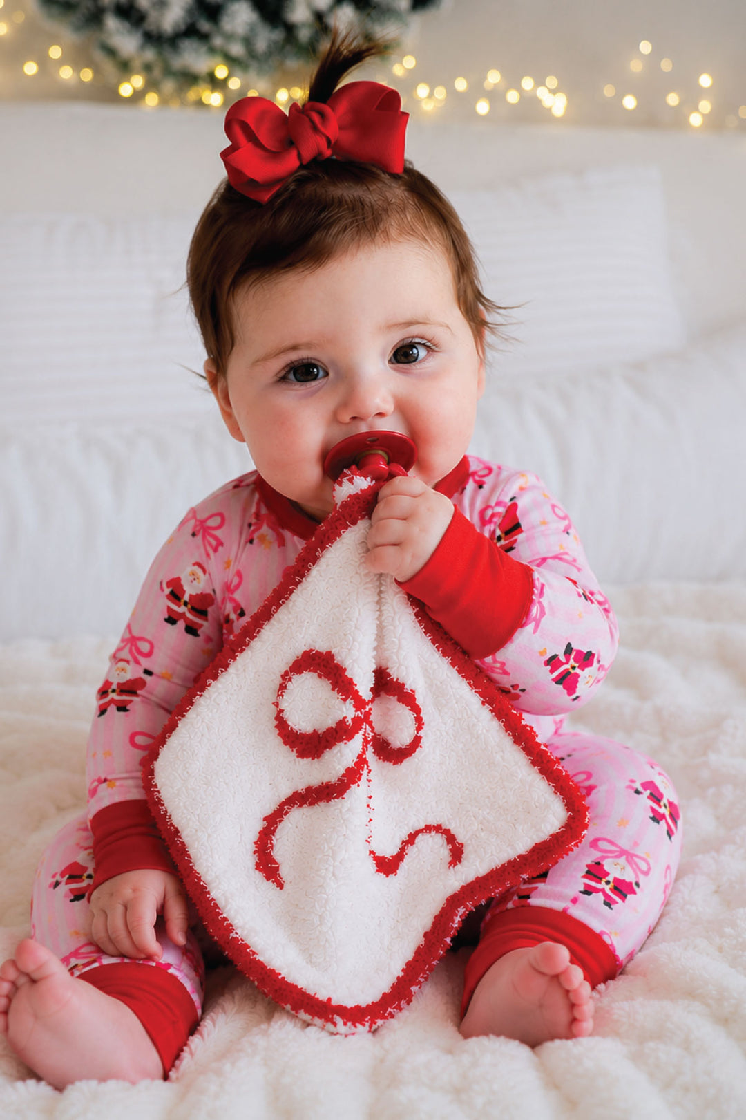 Baby in Caden Lane pink Santa print pajamas holding a red and white bow lovie security blanket with pacifier, crawling pose on cozy Christmas bedding