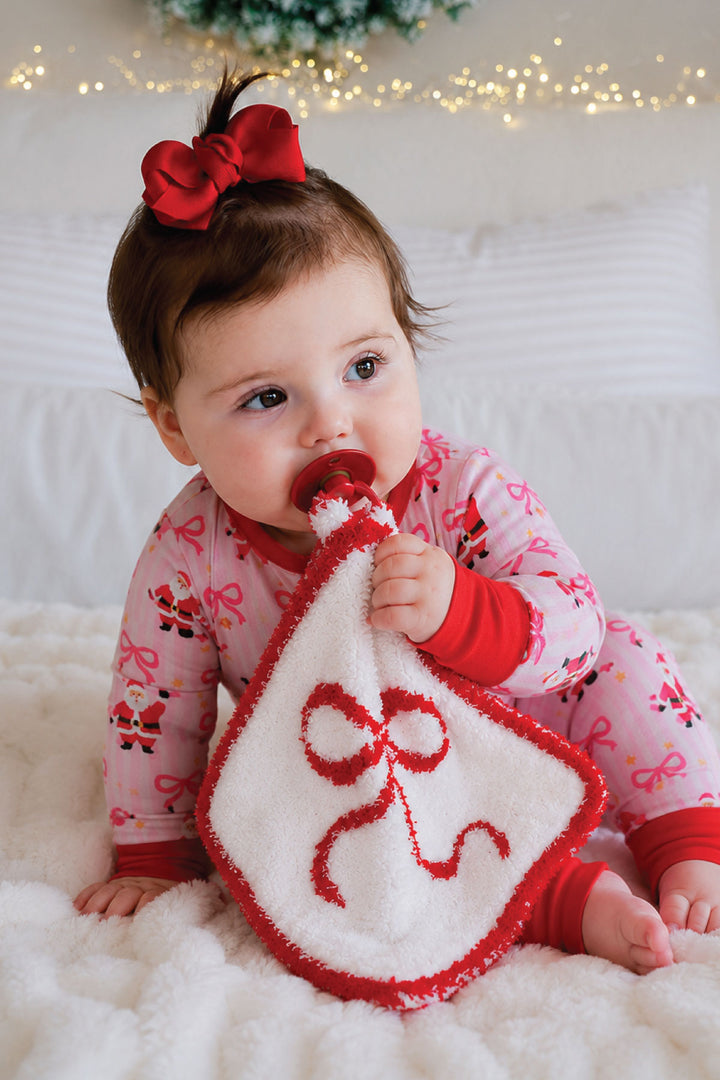 Baby girl in Caden Lane pink Santa and bow pajamas sitting on a bed with a red bow-embroidered lovie security blanket and pacifier, festive Christmas lights in background