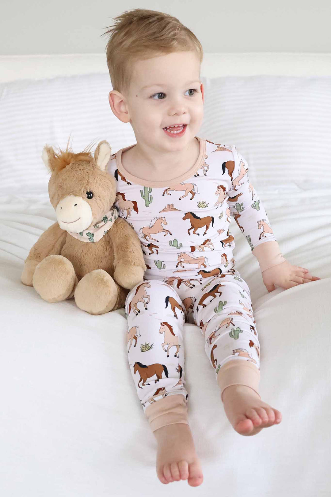 toddler boy sitting on bed in pony club neutral two piece pajama set next to stuffed horse 