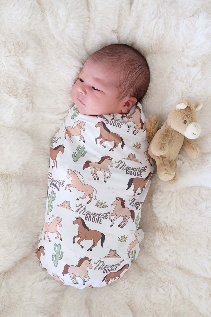 newborn boy wrapped up in pony club neutral personalized swaddle next to mini horse stuffed animal 