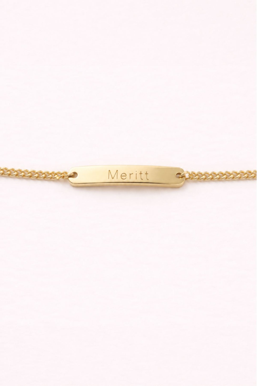 Caden Lane gold curb chain bracelet with engraved "Meritt" name bar charm on a soft pink background