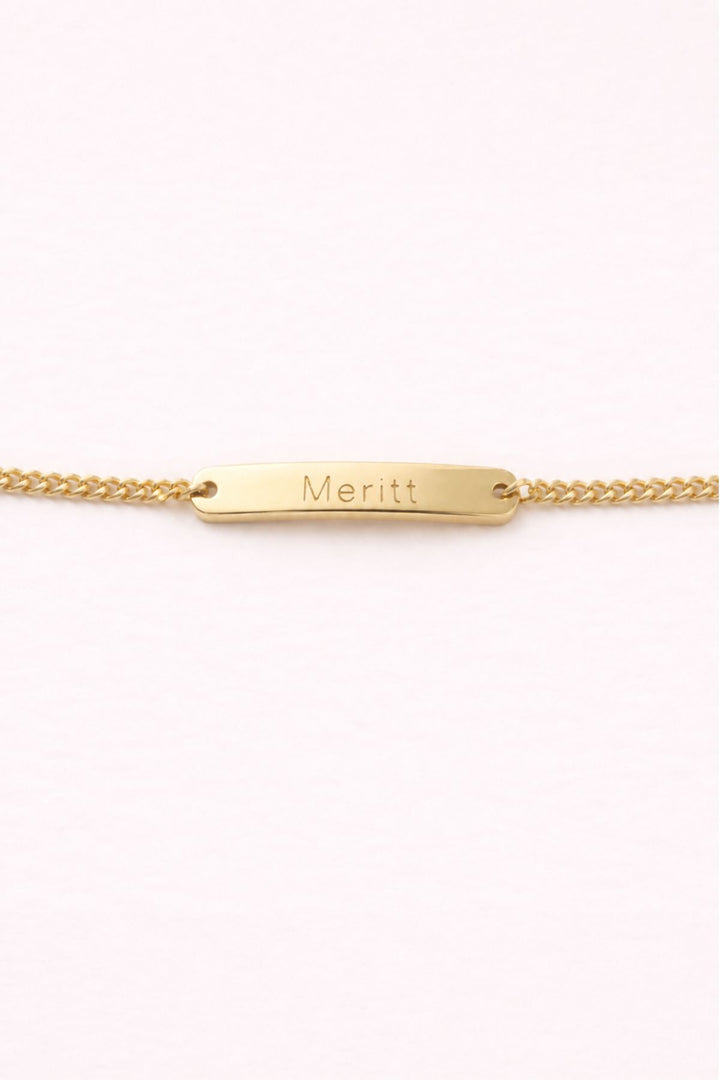 Caden Lane gold curb chain bracelet with engraved "Meritt" name bar charm on a soft pink background