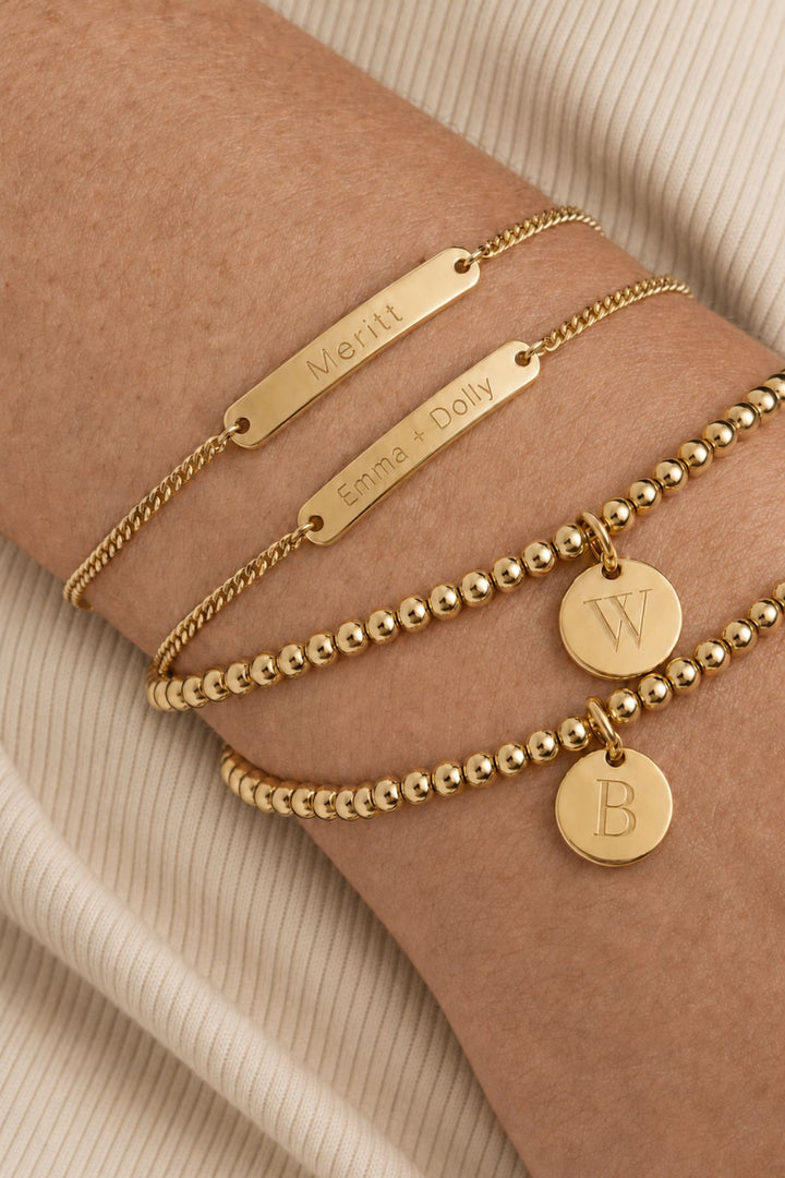 Caden Lane gold bracelet stack with engraved "Meritt" and "Emma + Dolly" name bars alongside beaded bracelets with initial discs "W" and "B"