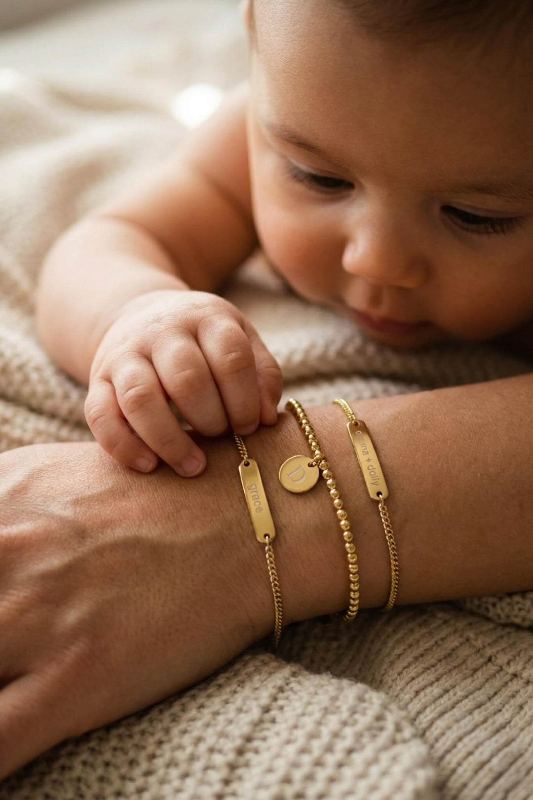 Caden Lane personalized gold bracelet stack with "Grace" name bar, initial "D" disc, and "Emma + Dolly" bar bracelet, baby reaching for mother's wrist