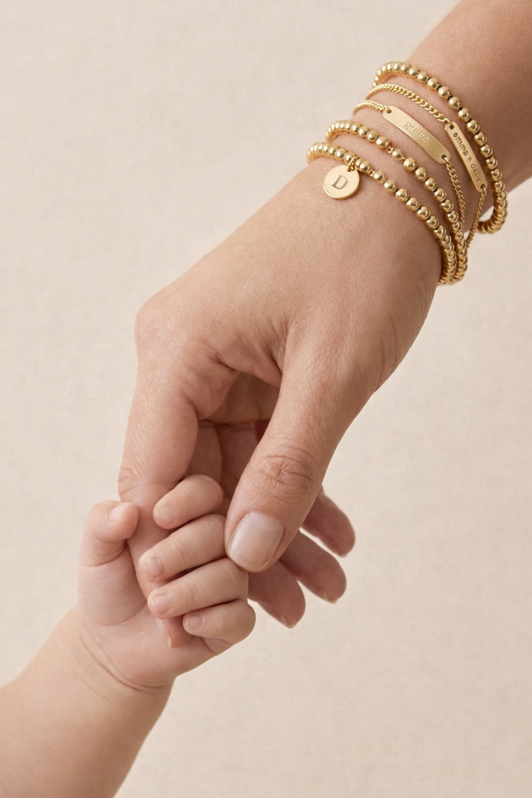 Caden Lane gold beaded bracelet stack with engraved bar and initial disc charms worn by a mother holding her baby's hand