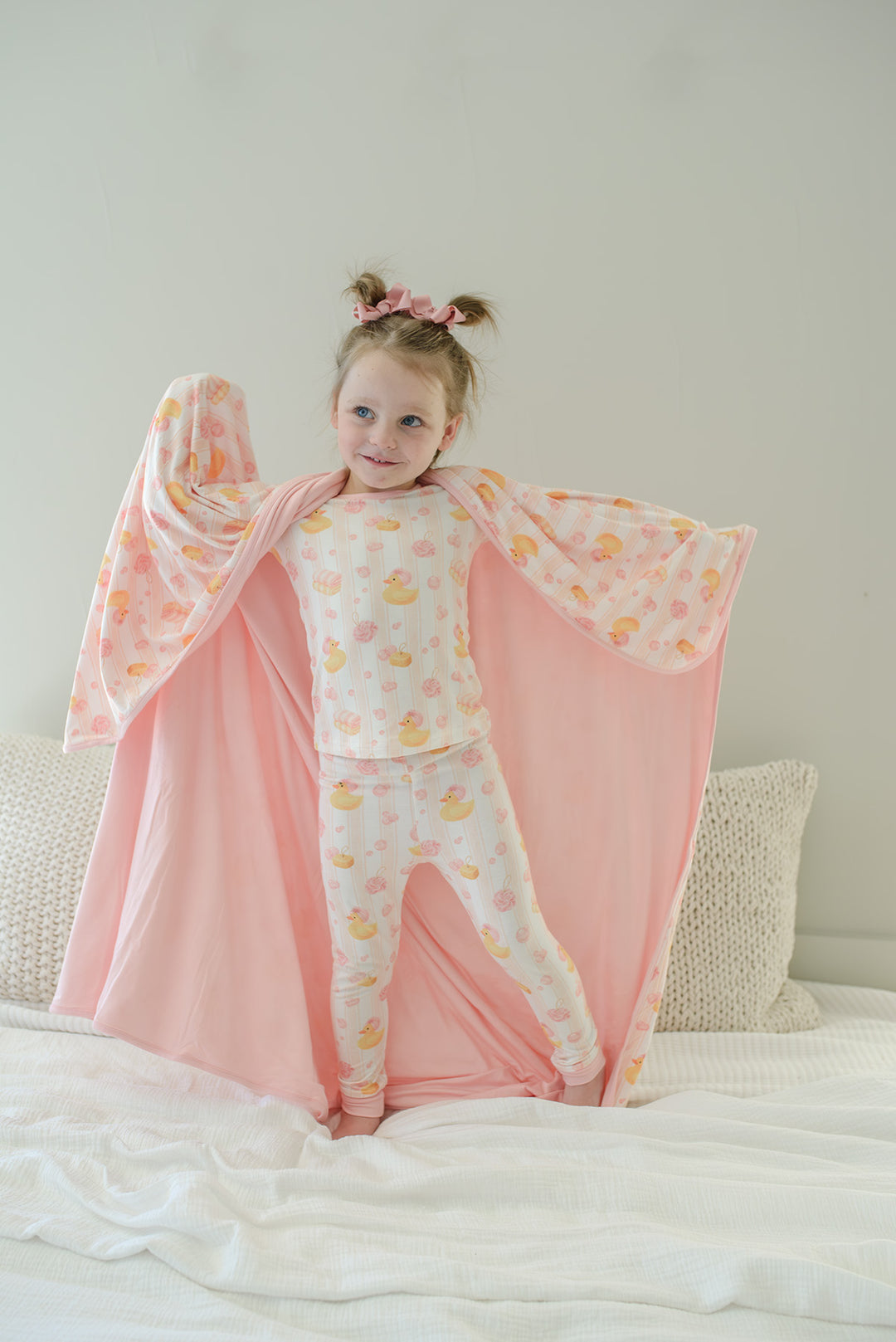 Young girl in matching pajamas holding wide open pink Bubble Duckies bamboo blanket showing printed and solid pink reverse sides