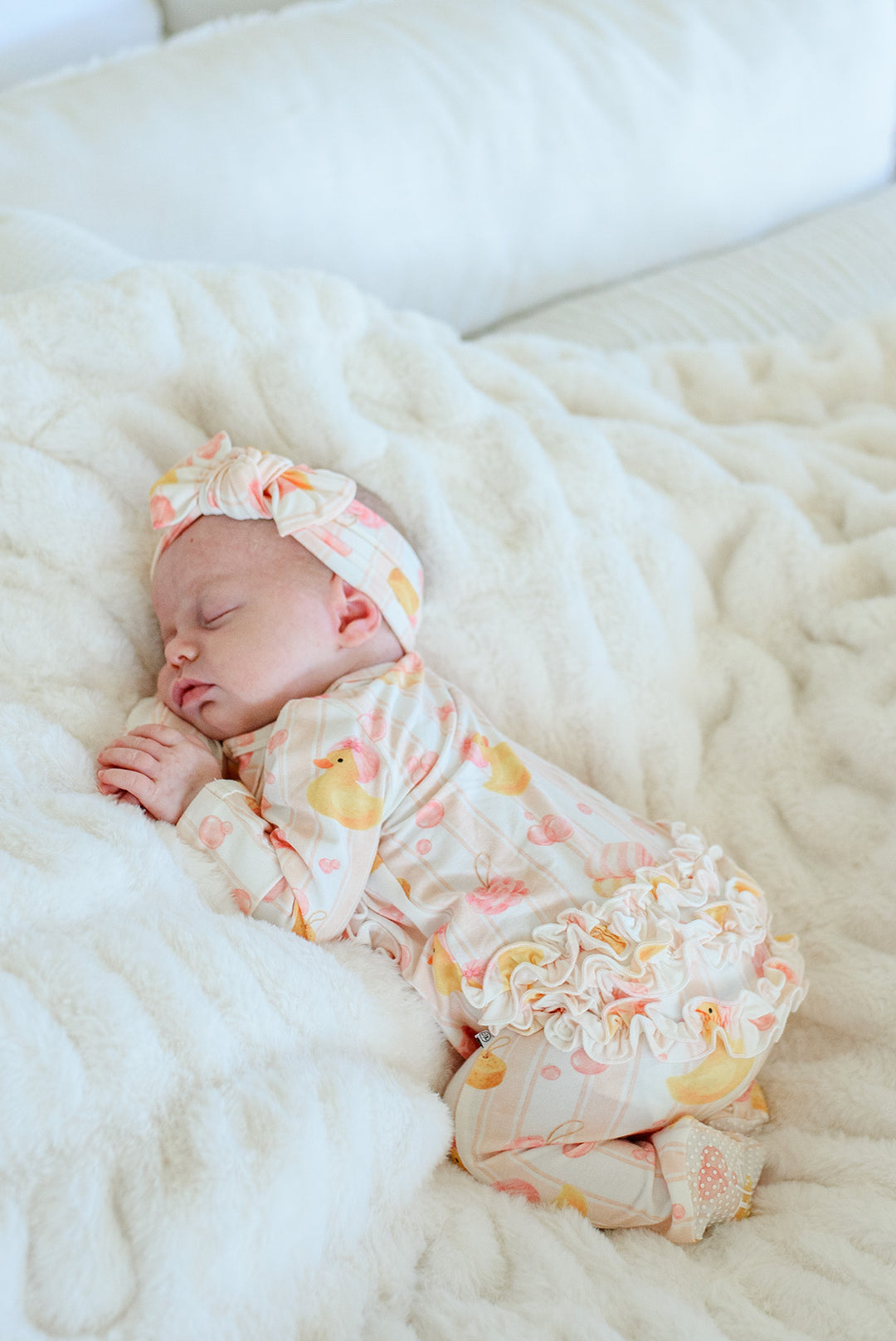 Sleeping newborn in pink Bubble Duckies ruffle zipper footie with bow headwrap on white fur blanket