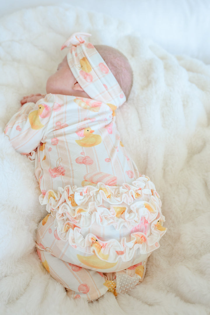 Newborn baby wearing pink Bubble Duckies ruffle footie pajamas and bow headwrap viewed from above on white fur