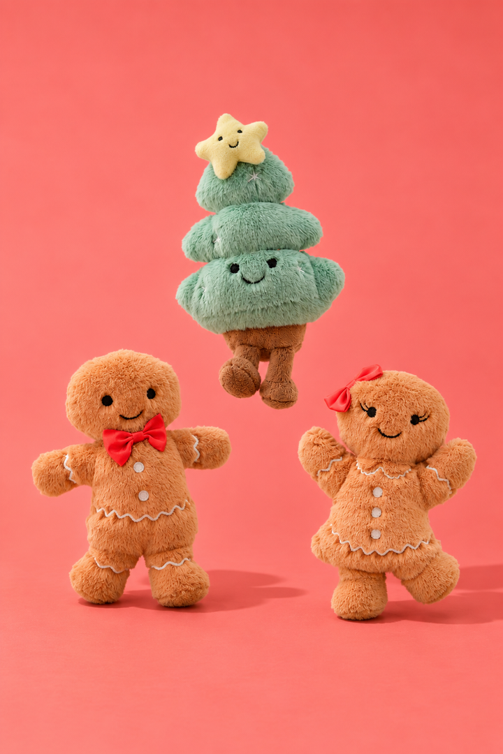 Caden Lane plush holiday toy set featuring a smiling green Christmas tree topped with a star, flanked by two gingerbread man plush toys, one with a red bow and one with a red bow tie, on a coral pink background.
