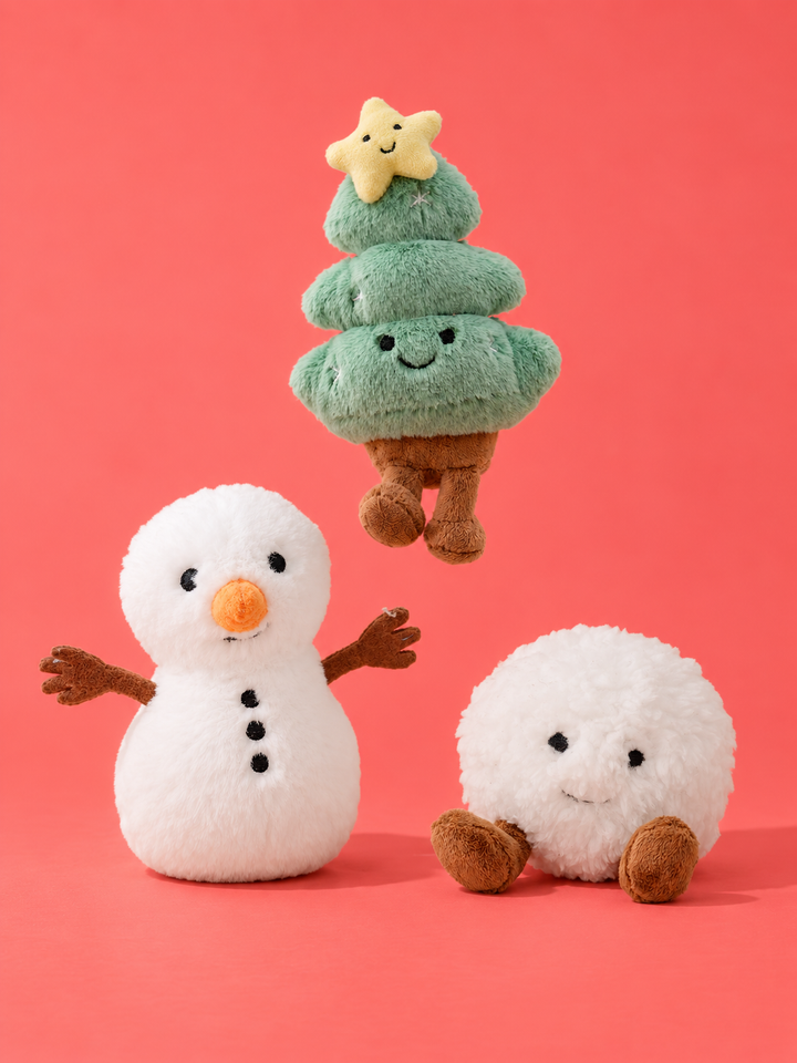 Caden Lane plush holiday toy set featuring a smiling snowman with a carrot nose, a green Christmas tree topped with a smiling star, and a round smiling snowball, all with brown stick-like arms and legs, on a coral pink background.