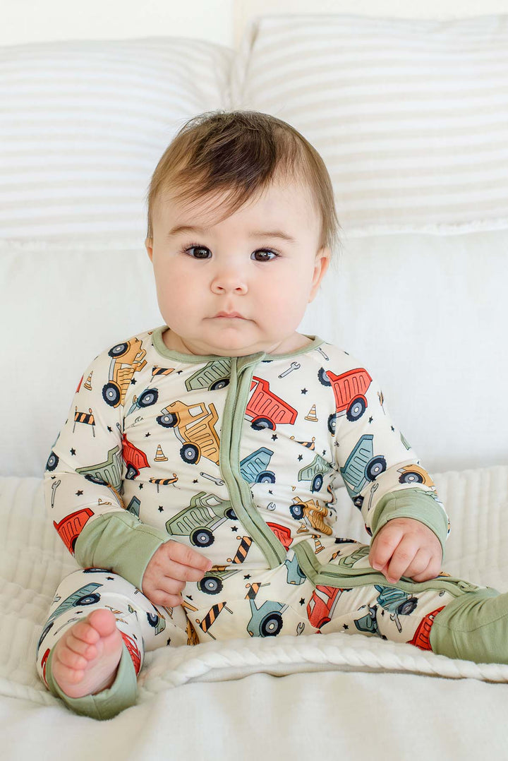 A brown-eyed baby sitting up against white pillows, wearing a Caden Lane cream knit zip romper in a Busy Builder print featuring dump trucks and cones, with sage trim.
