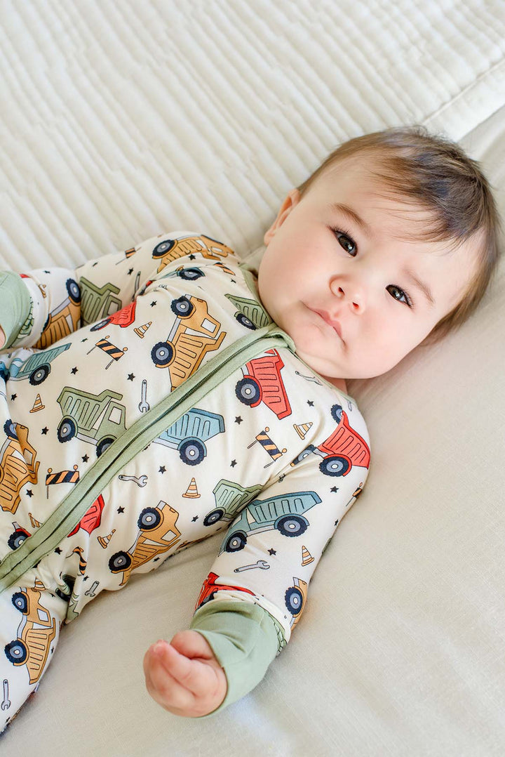 A baby lying on a cream quilt looking toward the camera, wearing a Caden Lane cream knit zip romper in a Busy Builder print featuring dump trucks and barricades.