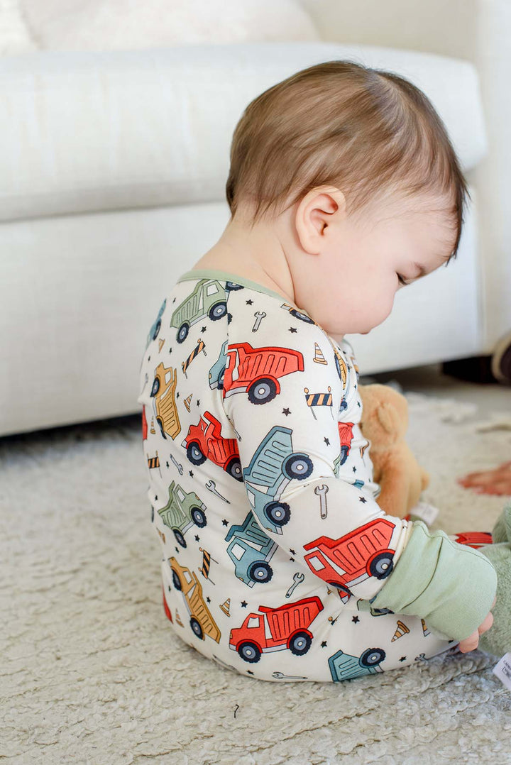 Back view of a baby sitting on a shag rug, wearing a Caden Lane cream knit romper in a Busy Builder print featuring dump trucks, paired with a teddy bear.
