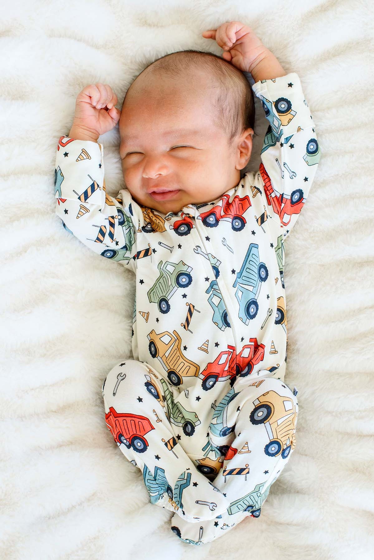 A smiling newborn stretching both arms overhead, eyes closed, wearing a Caden Lane cream knit footed sleeper in a Busy Builder print featuring dump trucks and cones.