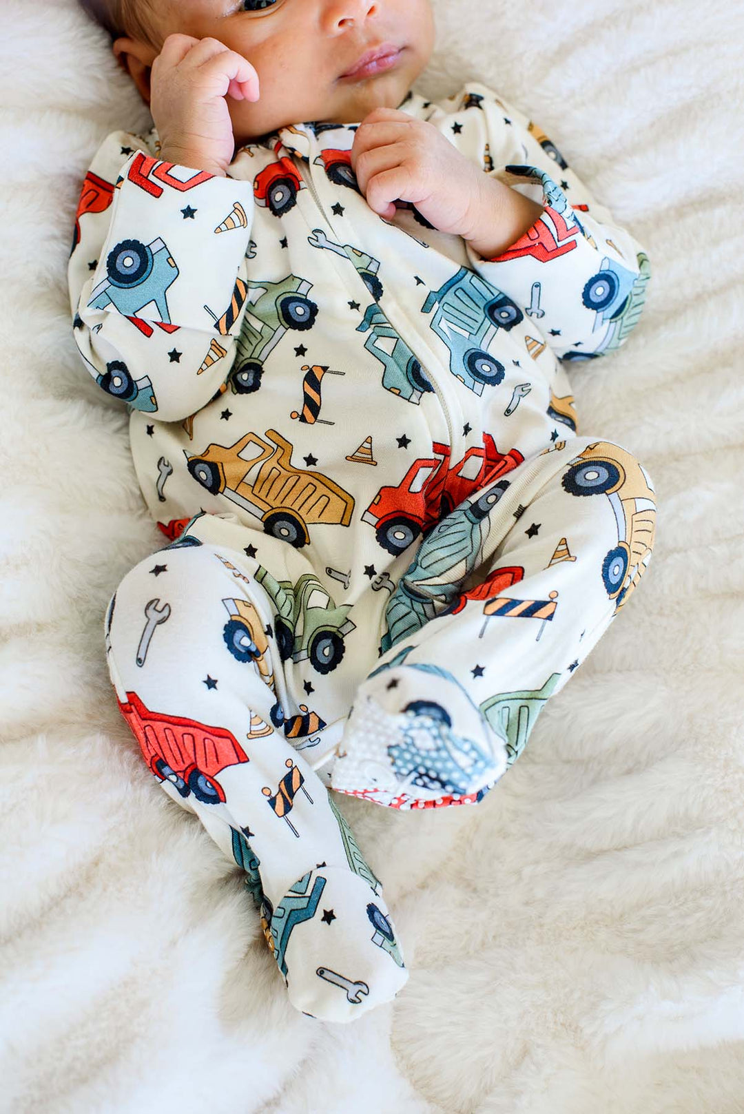 Cropped shot of a newborn with a hand by the cheek, wearing a Caden Lane cream knit footed sleeper in a Busy Builder print featuring dump trucks and barricades.