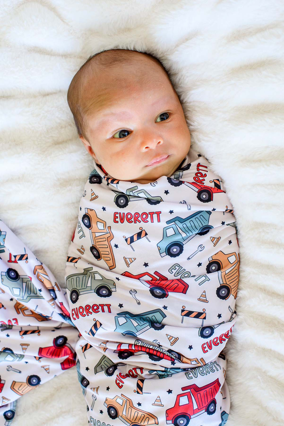 A newborn glancing sideways on white faux fur, wrapped in a Caden Lane cream swaddle in a Busy Builder print featuring dump trucks, reading "Everett."