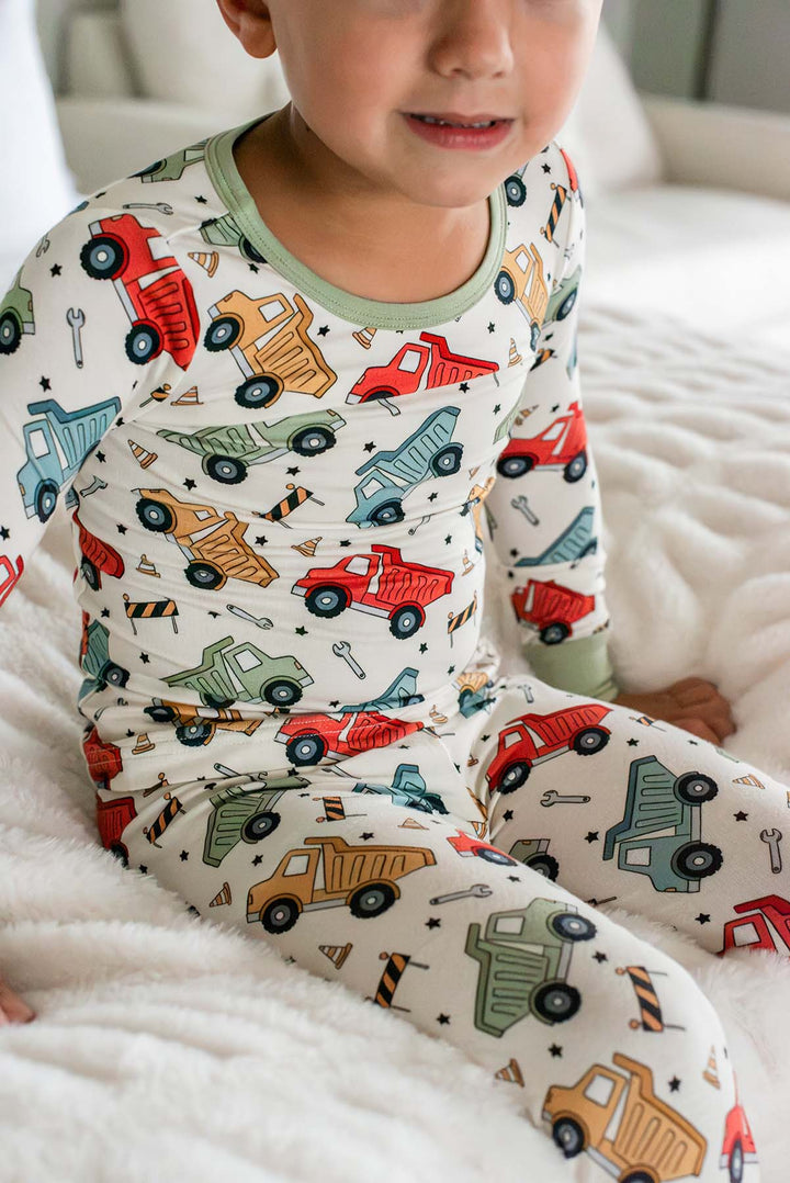 Cropped shot of a smiling toddler sitting on faux fur wearing a Caden Lane cream knit pajama set in a Busy Builder print featuring dump trucks, cones, and wrenches.