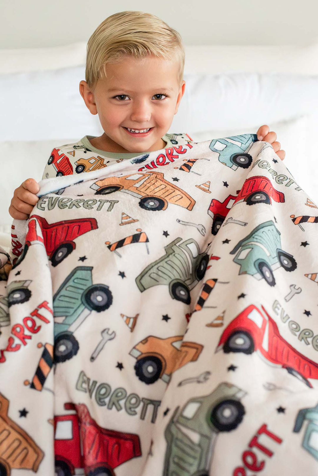 A grinning blond boy holding up a Caden Lane cream plush blanket in a Busy Builder print featuring dump trucks, cones, and wrenches, reading "Everett" in red and green.
