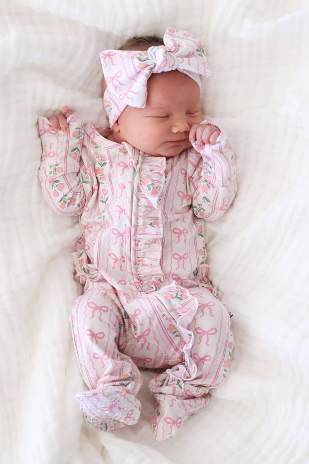 Newborn baby in a pink floral onesie and headband lying on a white blanket.
