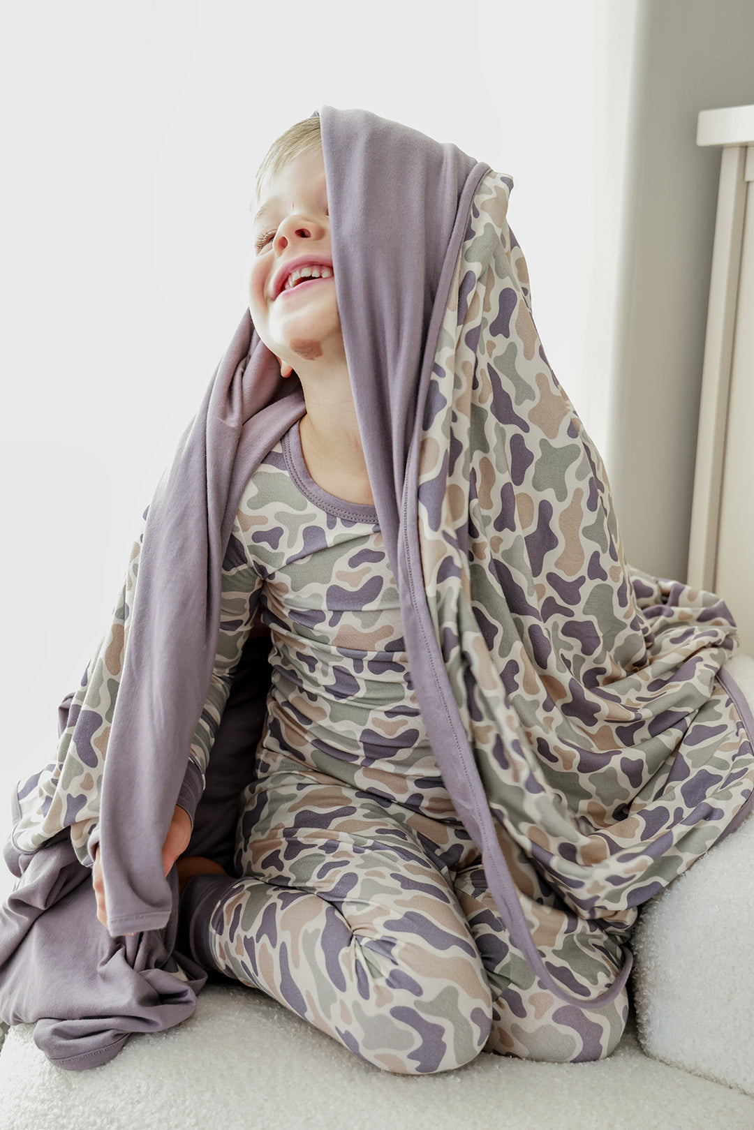 Smiling toddler boy with double-sided bamboo blanket draped over his head showing lavender interior, wearing matching camo cutie pajamas