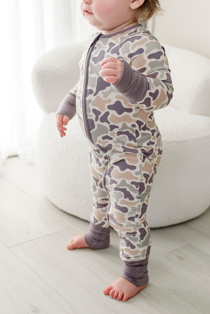 Toddler standing and showing side view of camo cutie convertible zip romper with gray cuffs