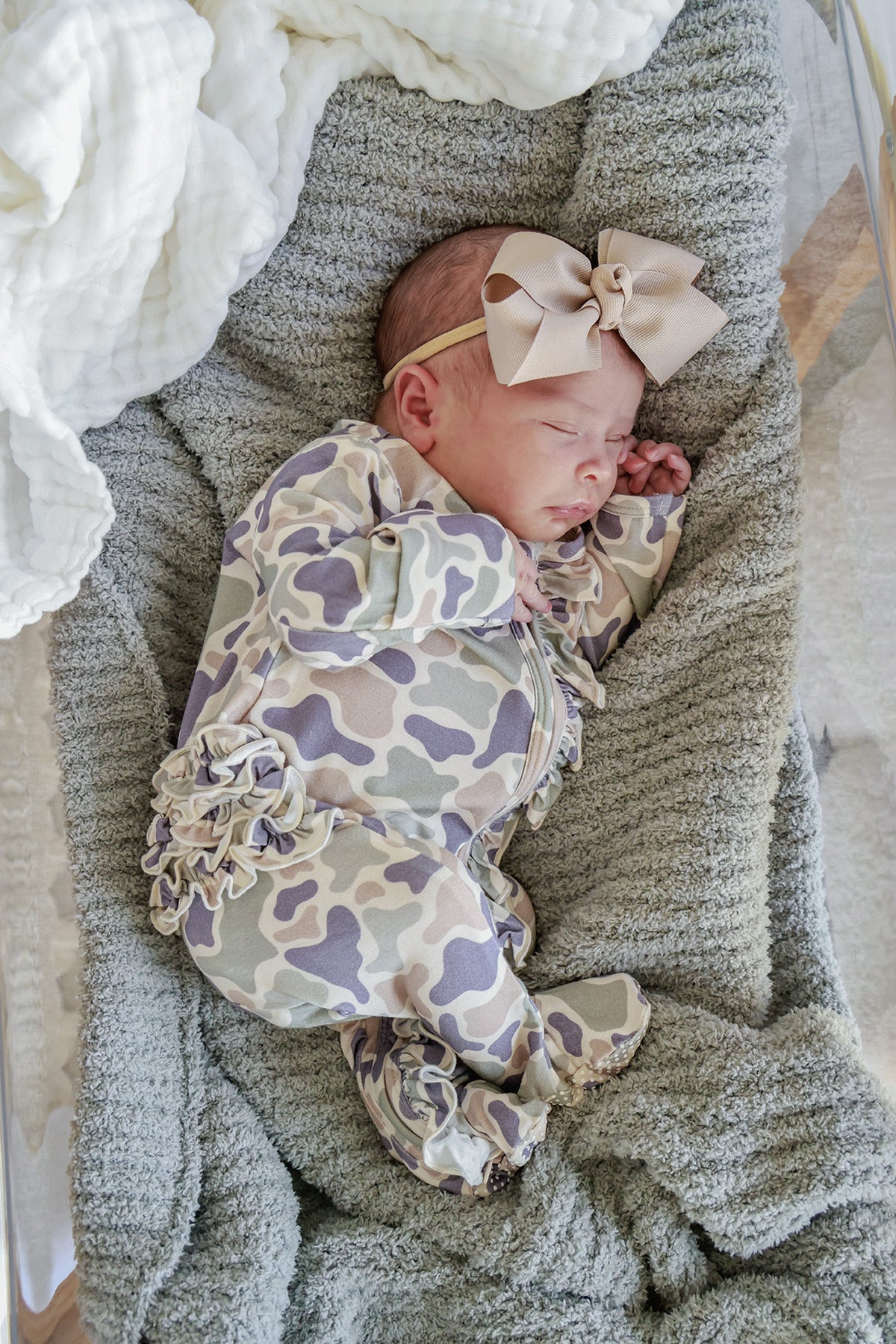 Newborn baby girl sleeping in a camo cutie print ruffle footie with a taupe ribbon bow headband on a gray knit blanket