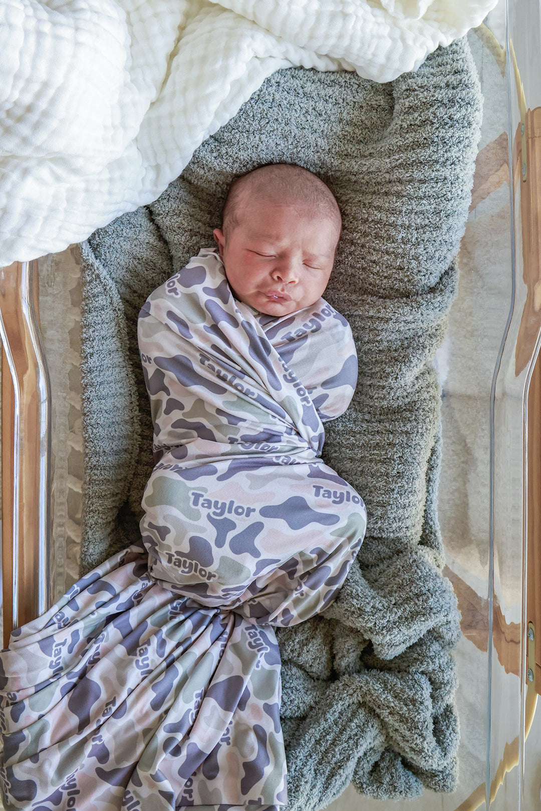 Newborn swaddled in a personalized camo cutie swaddle blanket with custom name, lying in a bassinet on a gray knit blanket
