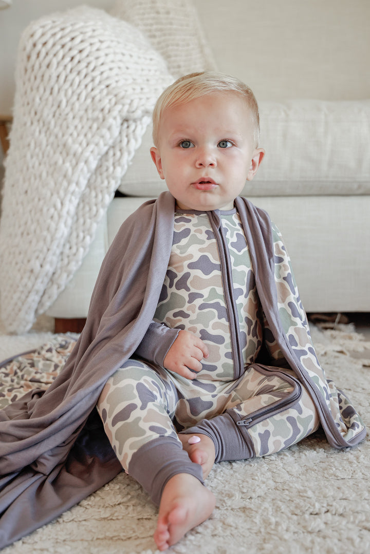 Back view of baby toddler in camo cutie convertible zip romper sitting on a rug