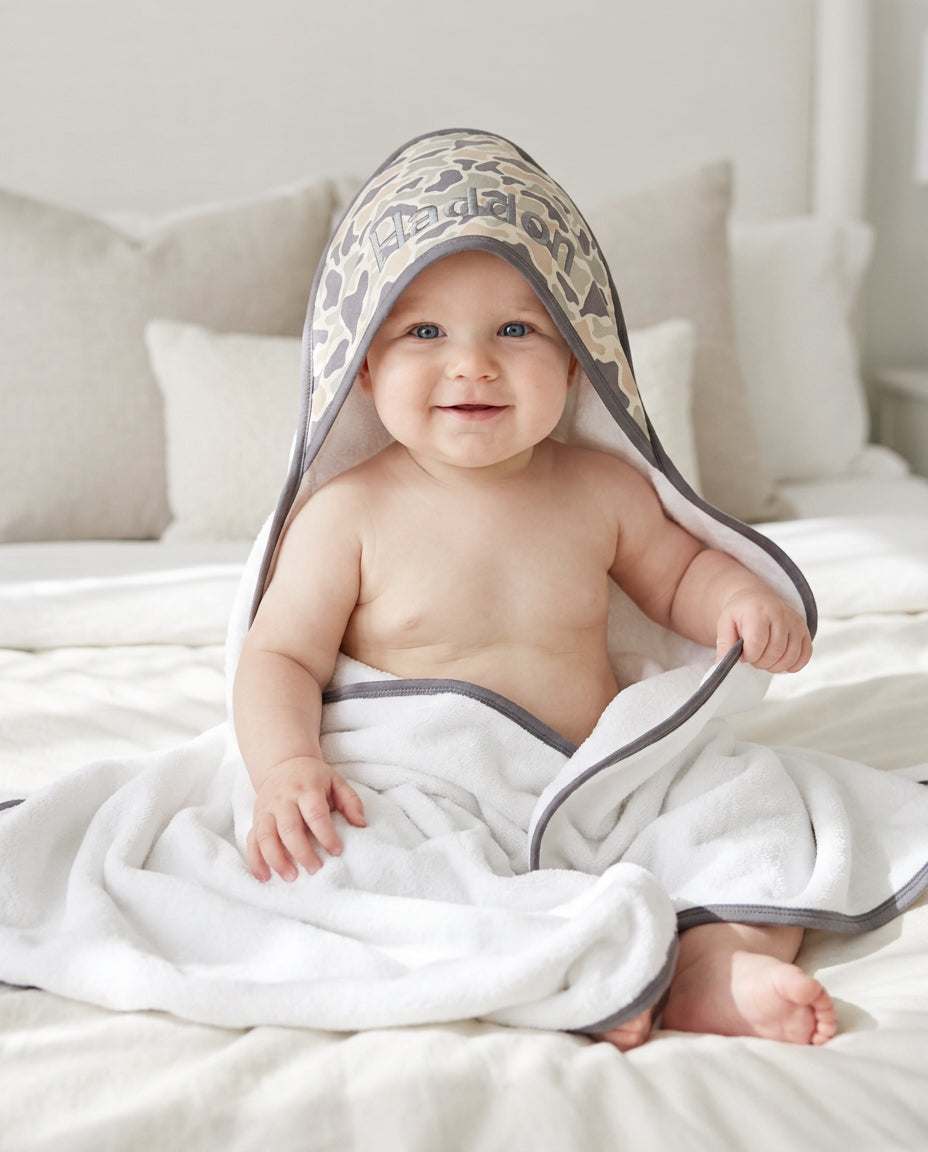 Baby sitting on bed wrapped in a bamboo hooded towel with a camo cutie animal print hood in tan and brown with gray trim